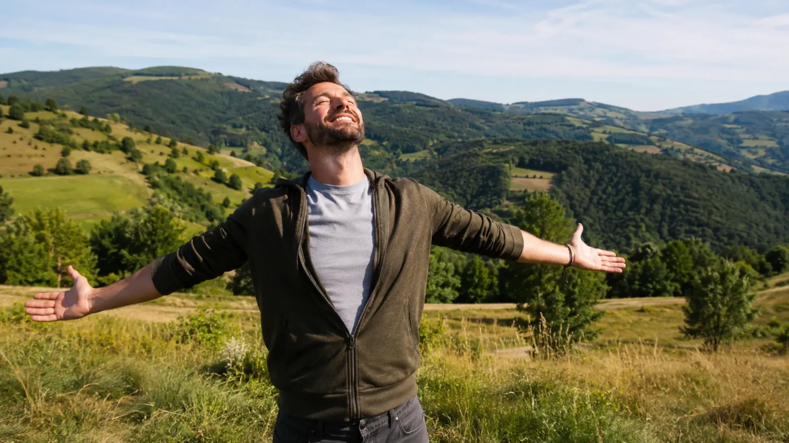 Homme souriant, bras ouverts, dans un paysage de collines verdoyantes